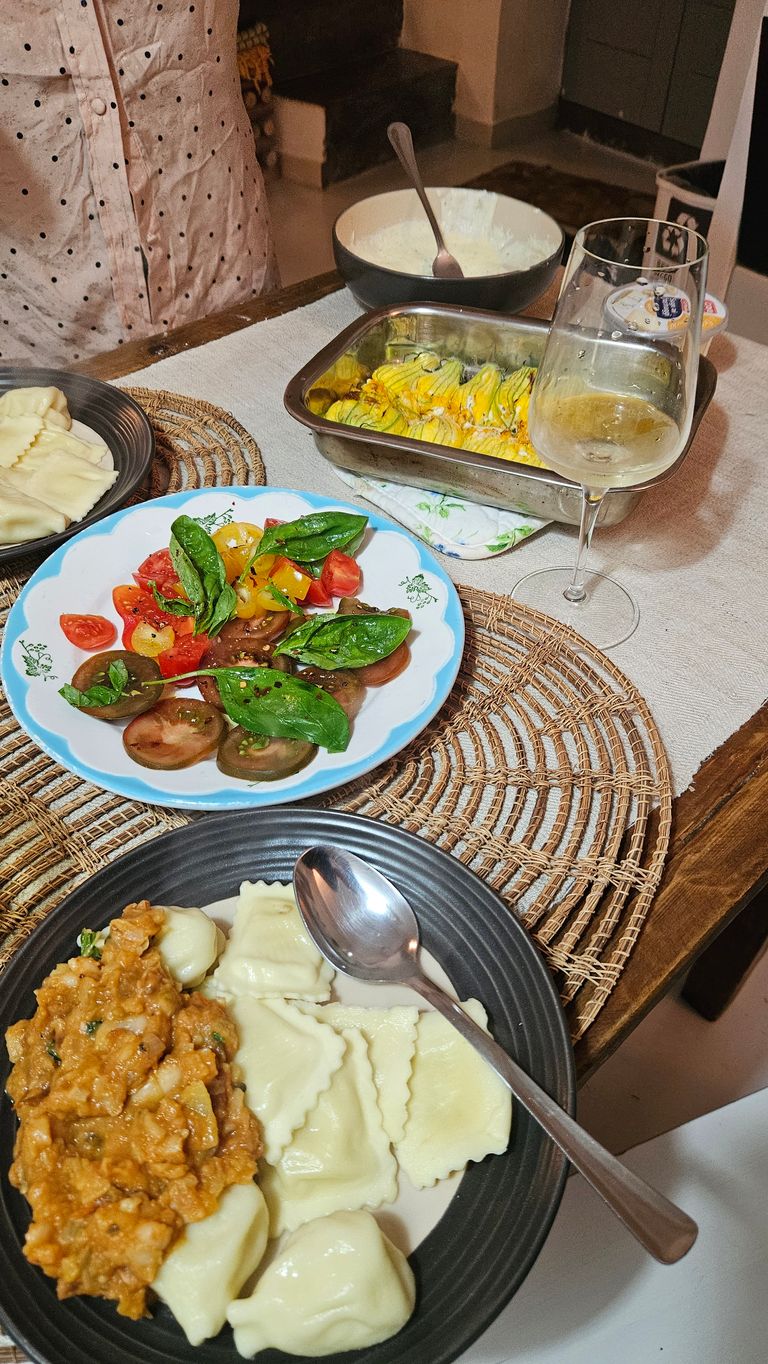 Auf einem gedeckten Tisch befinden sich zwei Teller mit Pasta, ein Teller mit Tomatensalat und ein Glas Weißwein. Im Hintergrund steht eine Person mit einem weißen, gepunkteten Hemd.