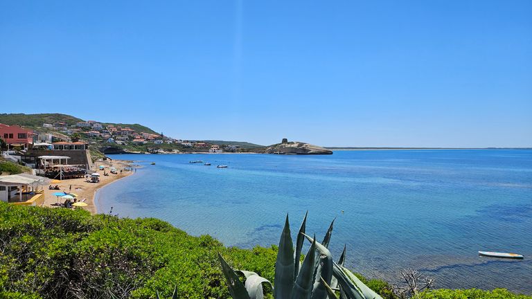 Blick auf eine Küste mit klarem, blauem Wasser, kleinen Booten und einem Sandstrand neben einer Wohnsiedlung unter blauem Himmel.