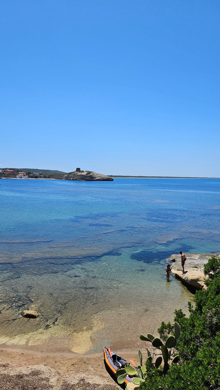 Zwei Personen an einem felsigen Ufer neben klarem, blauem Wasser unter einem wolkenlosen Himmel am Strand mit einem Kajak im Vordergrund und einer felsigen Insel im Hintergrund.