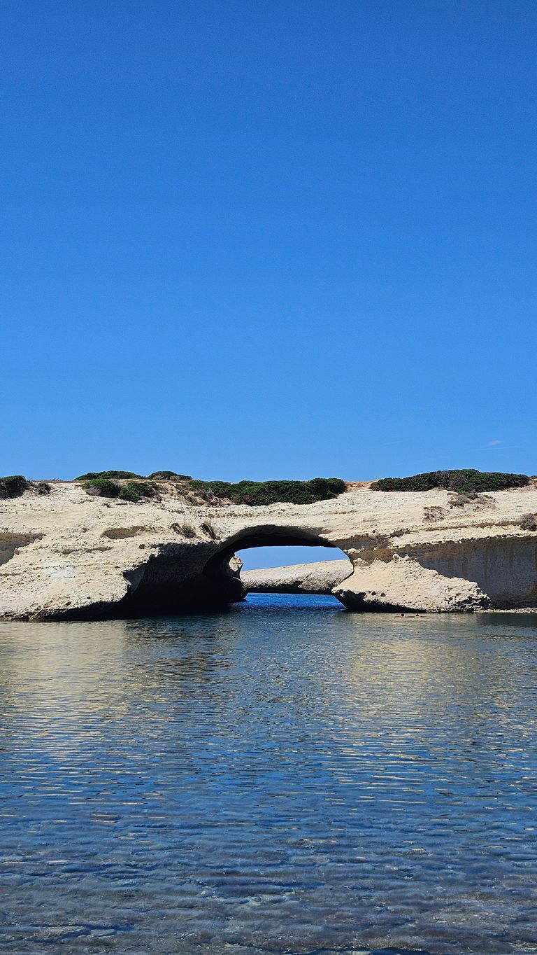 Ein natürliches Felsentor über klarem, ruhigem Wasser unter einem wolkenlosen blauen Himmel mit niedrigen grünen Pflanzen auf dem Felsplateau.