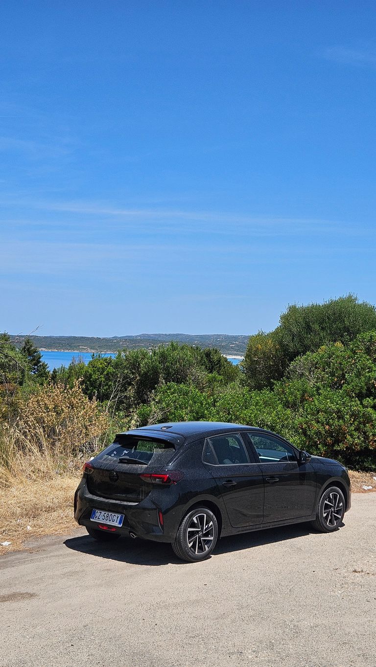 Schwarzes Auto, geparkt auf einem Asphaltweg, umgeben von grünem Gebüsch und trockenem Gras, mit Blick auf eine Küstenlandschaft unter blauem Himmel.