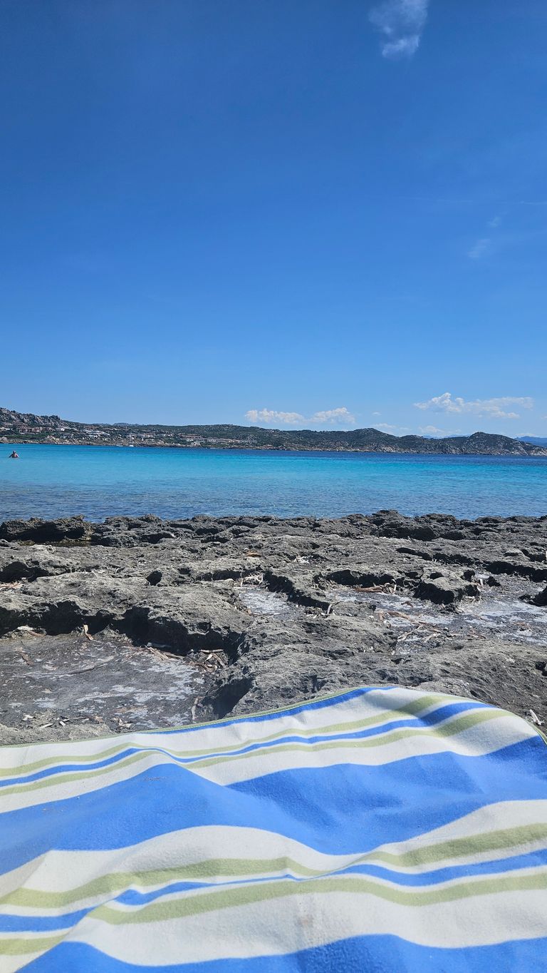 Felsen am Ufer mit Blick auf das klare, türkisfarbene Meer und einen teils bewachsenen Hügel im Hintergrund unter einem klaren blauen Himmel. Im Vordergrund liegt ein blau-grün gestreiftes Handtuch auf den Felsen.
