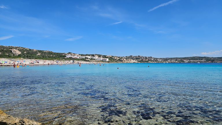 Klares türkisfarbenes Meer vor einem felsigen Strand mit Menschen im Wasser und weißen Gebäuden im Hintergrund unter blauem Himmel.