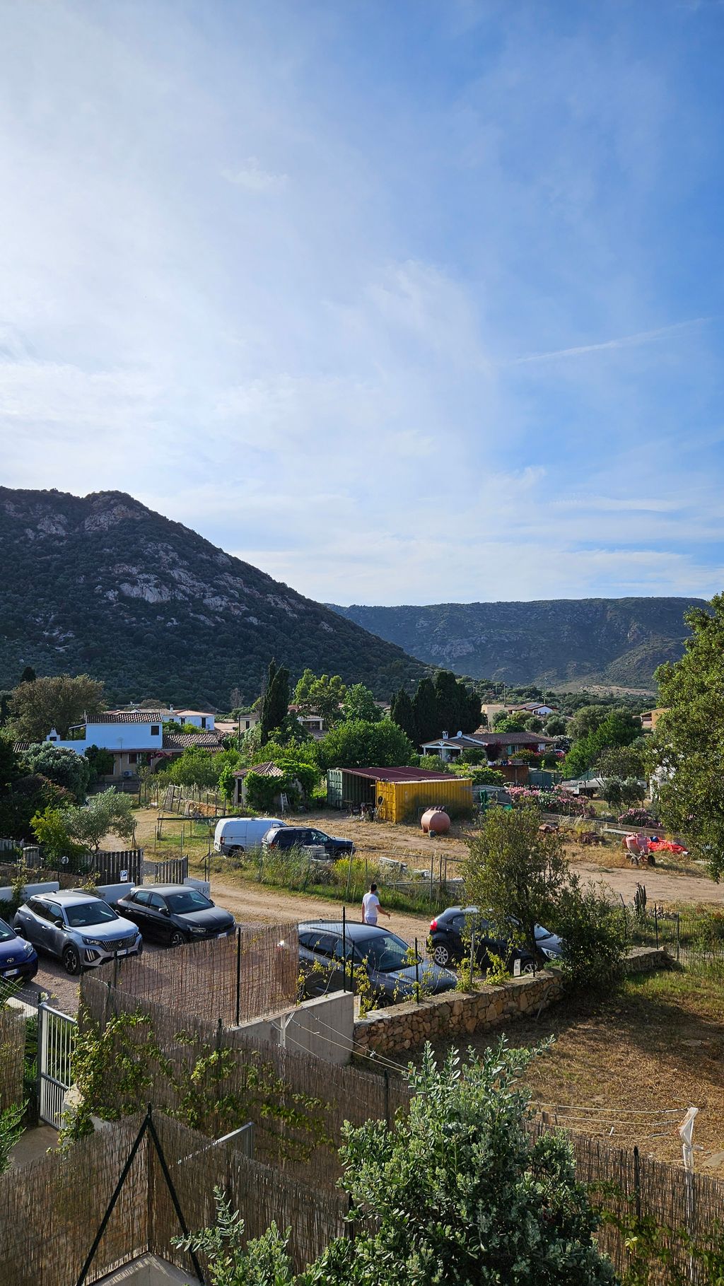Ländliche Landschaft mit Häusern, Autos auf einer dirt road, grünen Bäumen und Bergen im Hintergrund unter einem blauen Himmel mit Wolken.
