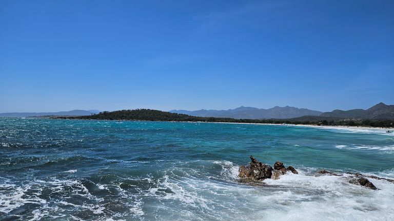 Klares blaues Meer mit Wellen, die gegen Felsen schlagen, im Vordergrund und eine bewaldete Küste mit Bergen im Hintergrund unter einem wolkenlosen blauen Himmel.