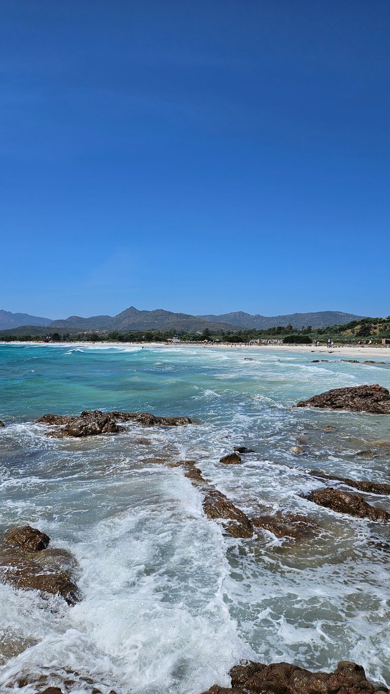 Wellen, die über Felsen an einem Strand mit Bergen im Hintergrund brechen, unter klarem blauem Himmel.