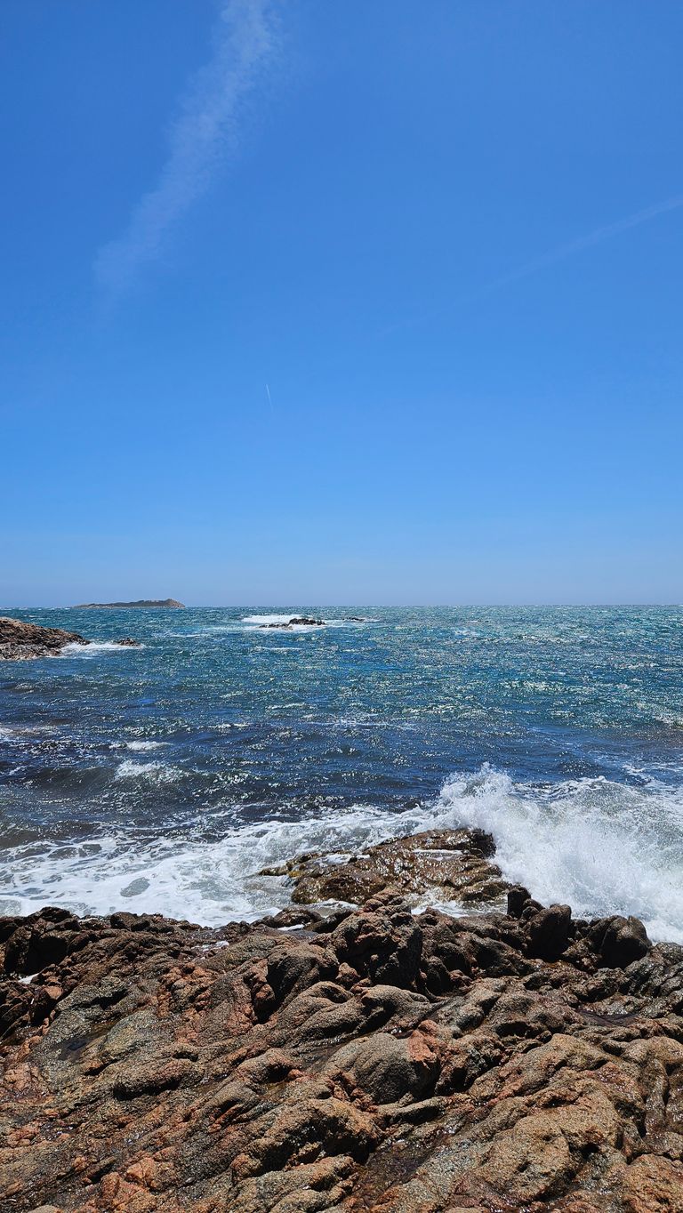 Krafter Küstenfelsen mit Wellen, die bei klarem Himmel ans Ufer schlagen, und Blick auf das offene Meer davor.