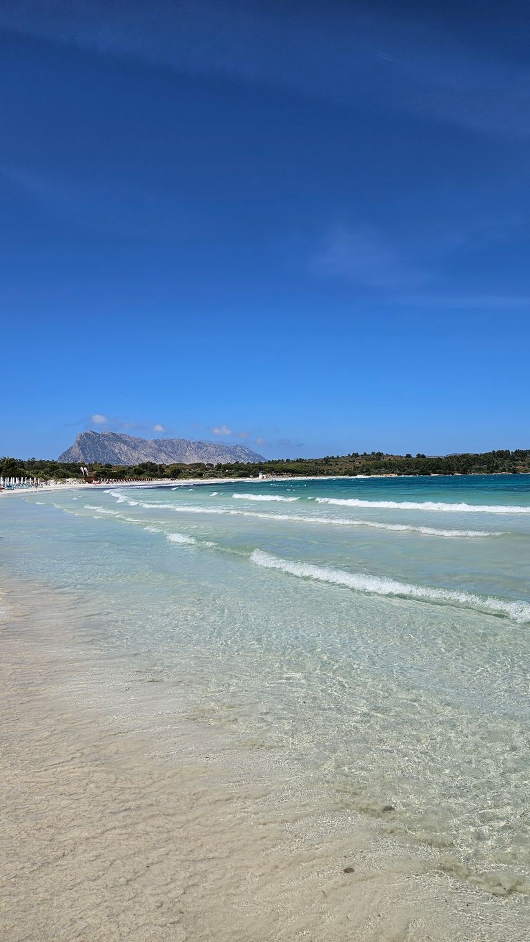 Klarer flacher Strand mit sanften Wellen und hellem Sand, im Hintergrund bewaldete Küste und ein großer Berg unter strahlend blauem Himmel.