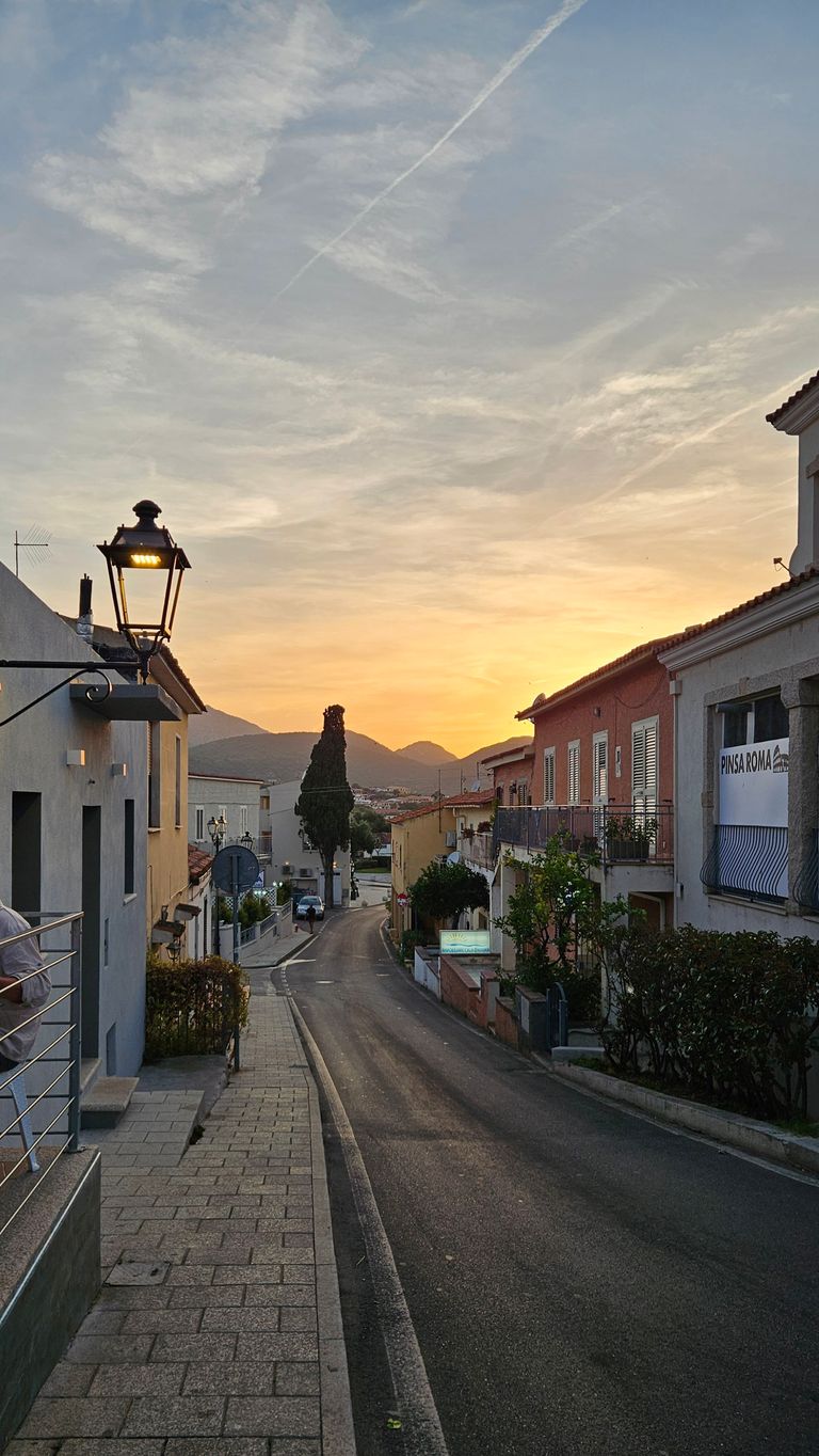 Eine schmale Straße in einem ruhigen Stadtviertel bei Sonnenuntergang mit Häusern auf beiden Seiten, einer Straßenlaterne und Bergen im Hintergrund.