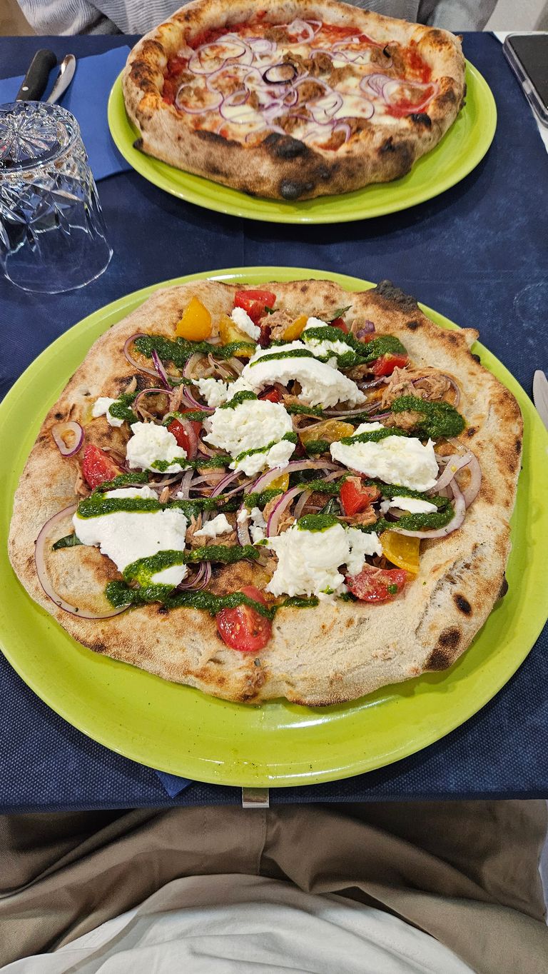 Zwei Pizzen auf grünen Tellern auf einem dunklen Tisch. Die vordere Pizza ist mit Frischkäse, roten und gelben Tomaten, roten Zwiebeln und Pesto belegt. Die hintere Pizza hat Mozzarella, rote Zwiebeln und Tomatensauce.