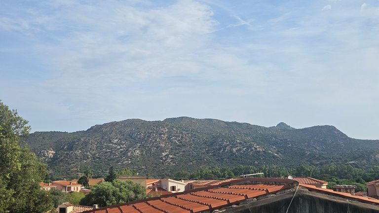 Blick auf ein Dorf mit roten Ziegeldächern vor bewaldeten Bergen unter einem leicht bewölkten Himmel.