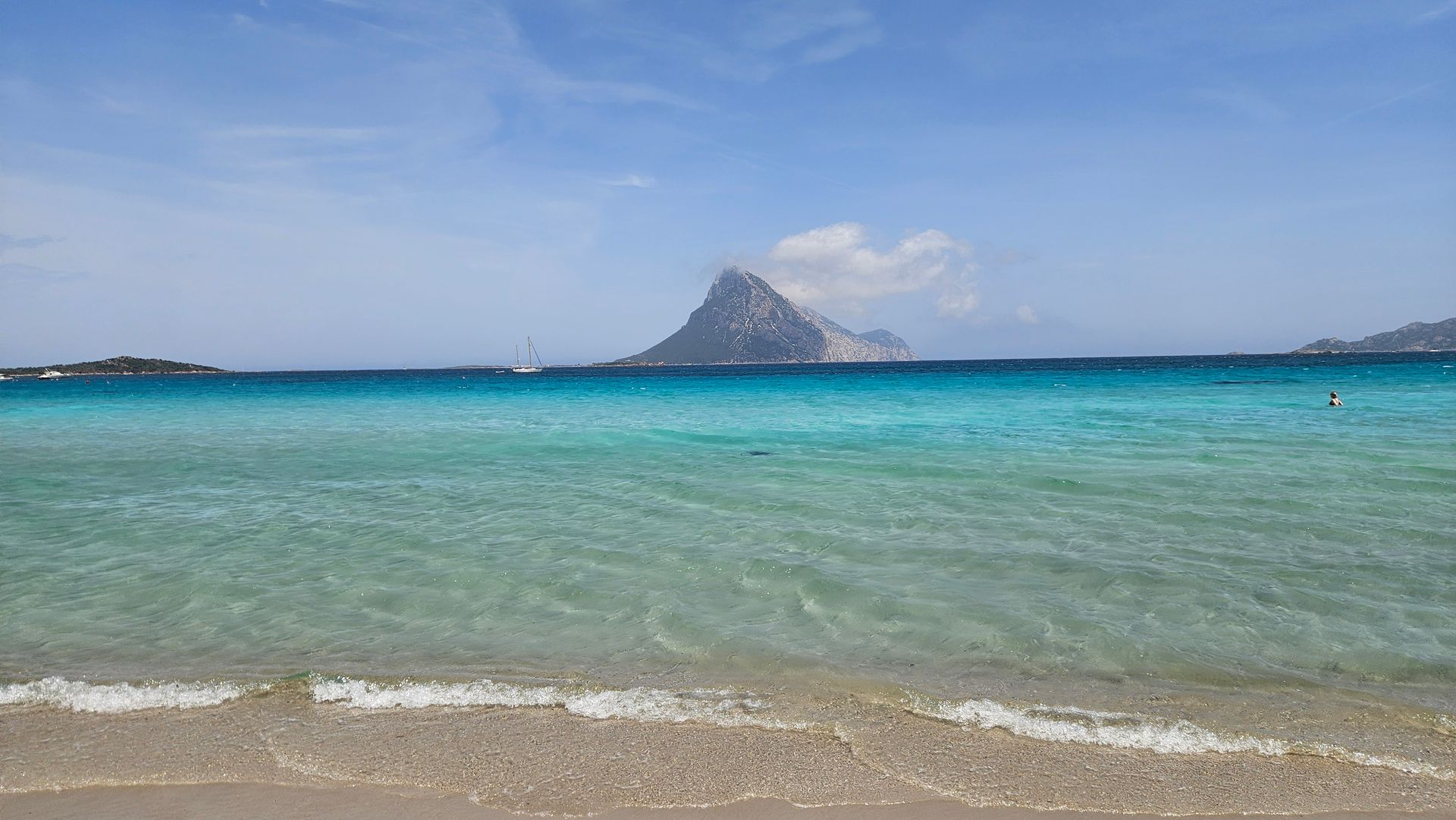 Klares türkisfarbenes Meer mit Sandstrand im Vordergrund und einer Insel mit bergigem Gipfel am Horizont unter blauem Himmel.