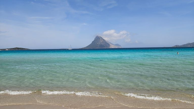 Klares türkisfarbenes Meer mit Sandstrand im Vordergrund und einer Insel mit bergigem Gipfel am Horizont unter blauem Himmel.