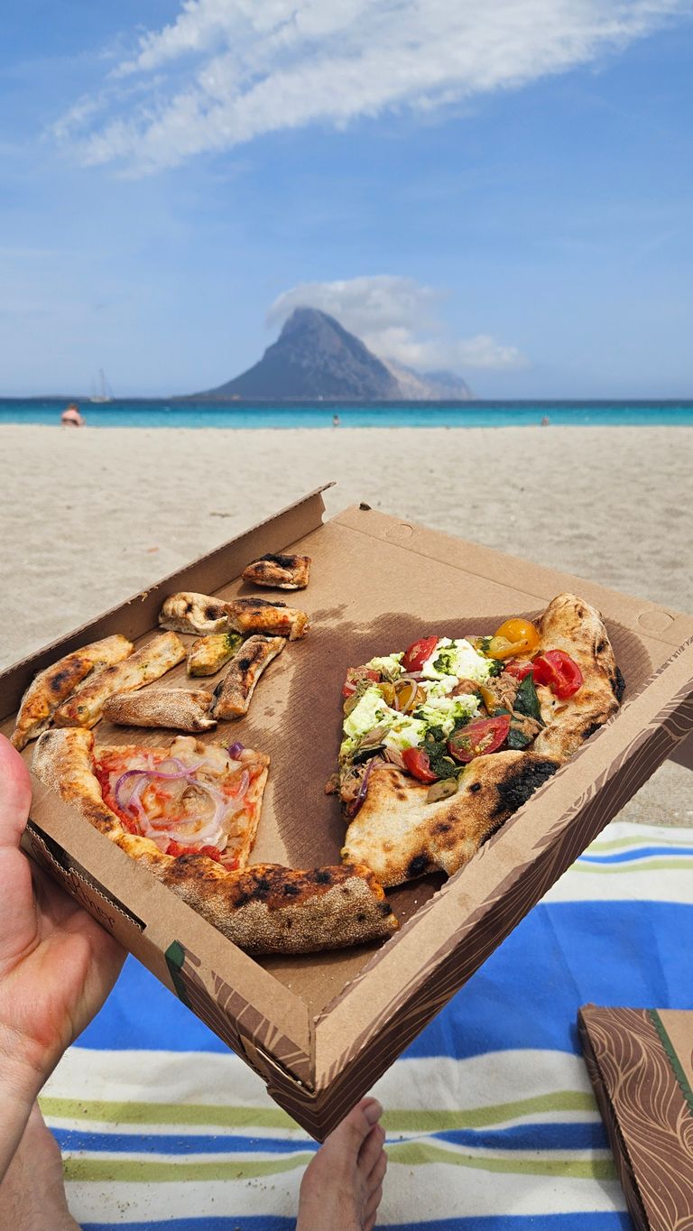 Eine teilweise aufgegessene Pizza in einer Pappschachtel wird an einem Sandstrand mit blauem Himmel und einer Insel im Hintergrund gehalten.