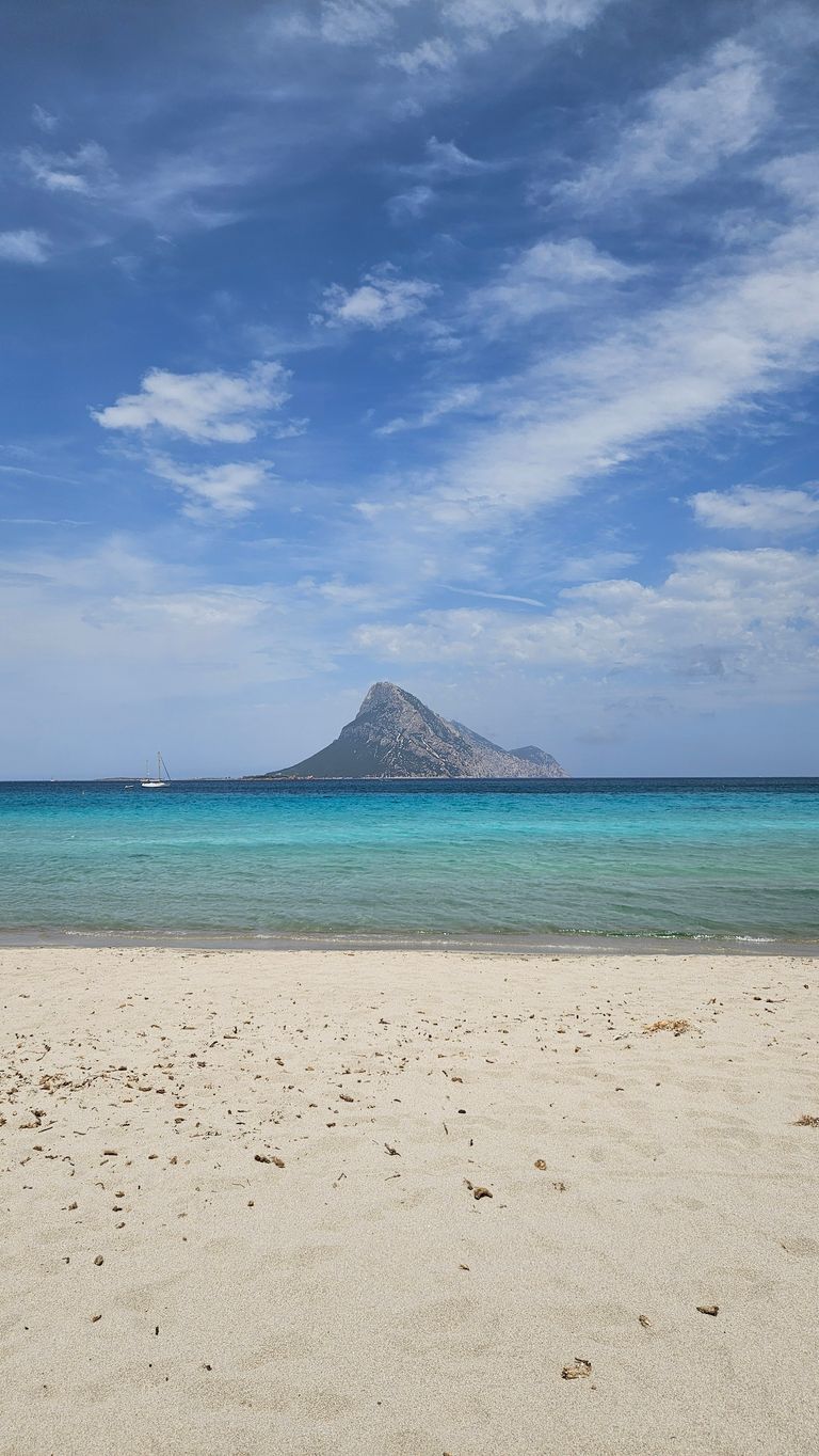 Ein ruhiger Sandstrand mit verstreuten kleinen Steinen im Vordergrund, türkisblaues Meer und eine felsige Insel unter einem teils bewölkten blauen Himmel im Hintergrund.