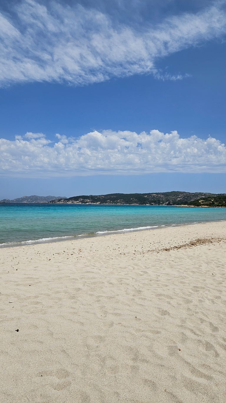 Ein weiter Sandstrand mit ruhigem, türkisfarbenem Wasser und einer felsigen Insel im Hintergrund unter blauem Himmel mit einigen Wolken.
