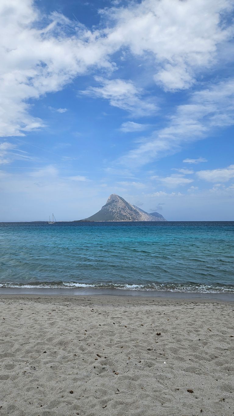 Ein einsamer Berg ragt aus dem Meer auf, davor ein Sandstrand und dahinter ein bewölkter blauer Himmel.
