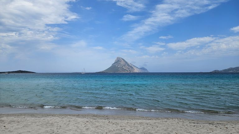 Strand mit Sand im Vordergrund, ruhigem türkisblauem Meer und einem markanten Berg im Hintergrund unter einem bewölkten Himmel.