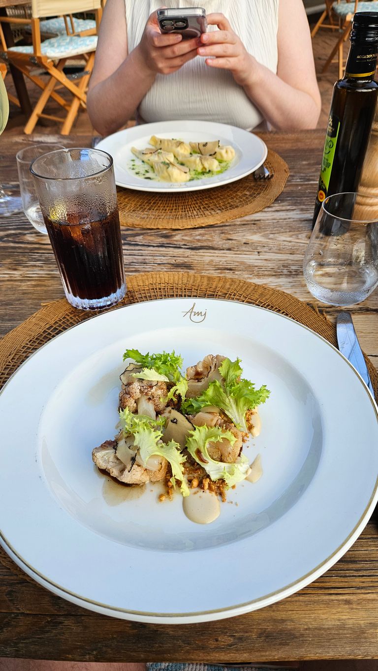 Zwei Teller mit elegant angerichteten Gerichten auf einem Holztisch in einem Restaurant, im Vordergrund ein Gericht mit Blumenkohl, Blattsalat und Soße, im Hintergrund eine Person, die ein Smartphone hält.