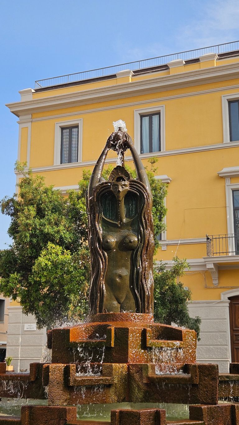 Bronzefigur einer Frau mit erhobenen Armen und Wasser, das aus ihren Händen sprudelt, in einem quadratischen steinernen Brunnen vor einem gelben Wohngebäude mit Bäumen im Hintergrund.