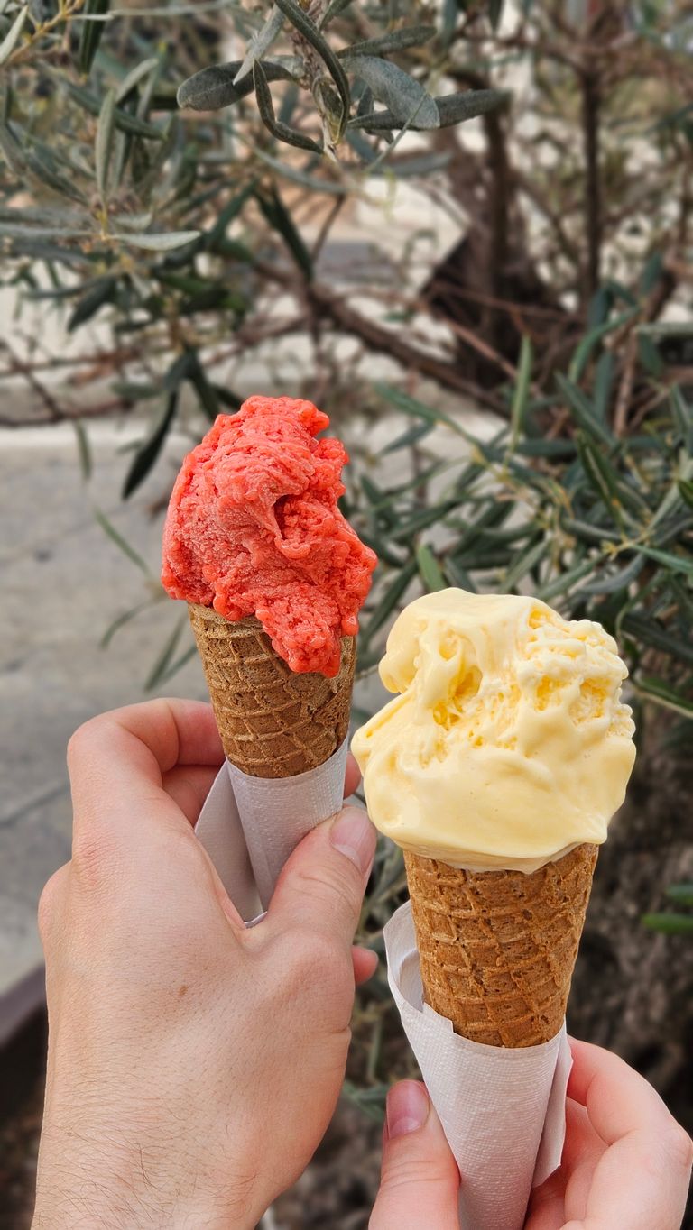 Zwei Hände halten zwei Eiswaffeln, eine mit rotem und die andere mit gelbem Eis, vor einem Hintergrund aus Zweigen und Blättern.