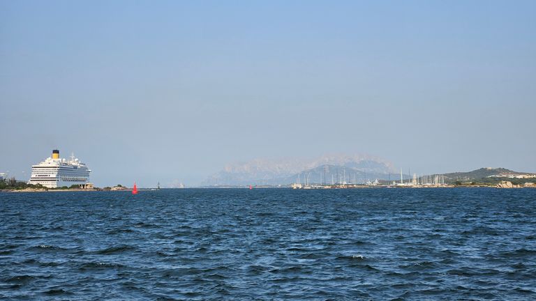 Ein Kreuzfahrtschiff liegt vor der Küste in der Ferne, mit Segelbooten und Hügeln im Hintergrund unter klarem Himmel.