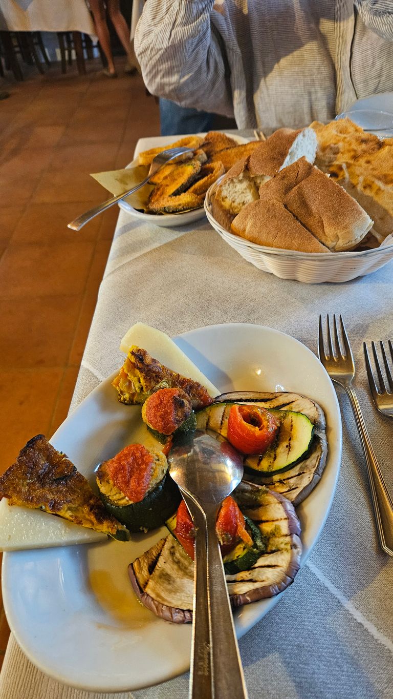 Teller mit gegrilltem Gemüse, gefüllten Zucchini und kleinen Frittata-Stücken, daneben ein Brotkorb mit verschiedenen Brotsorten auf einem Tisch mit Tischdecke in einem Restaurant.