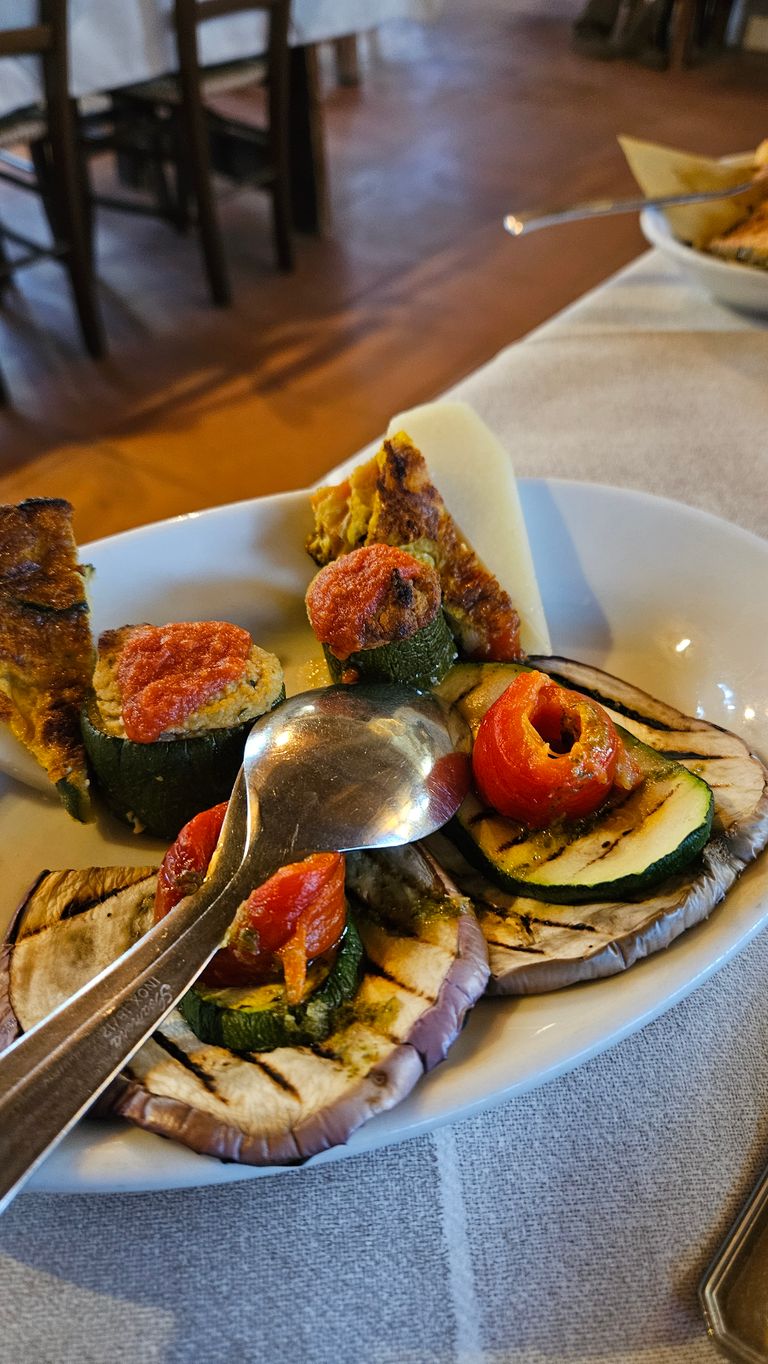 Ein weißer Teller mit gegrillten Auberginenscheiben, Zucchini mit Tomatensauce gefüllt und zwei Löffeln, auf einem gedeckten Tisch in einem Restaurant.