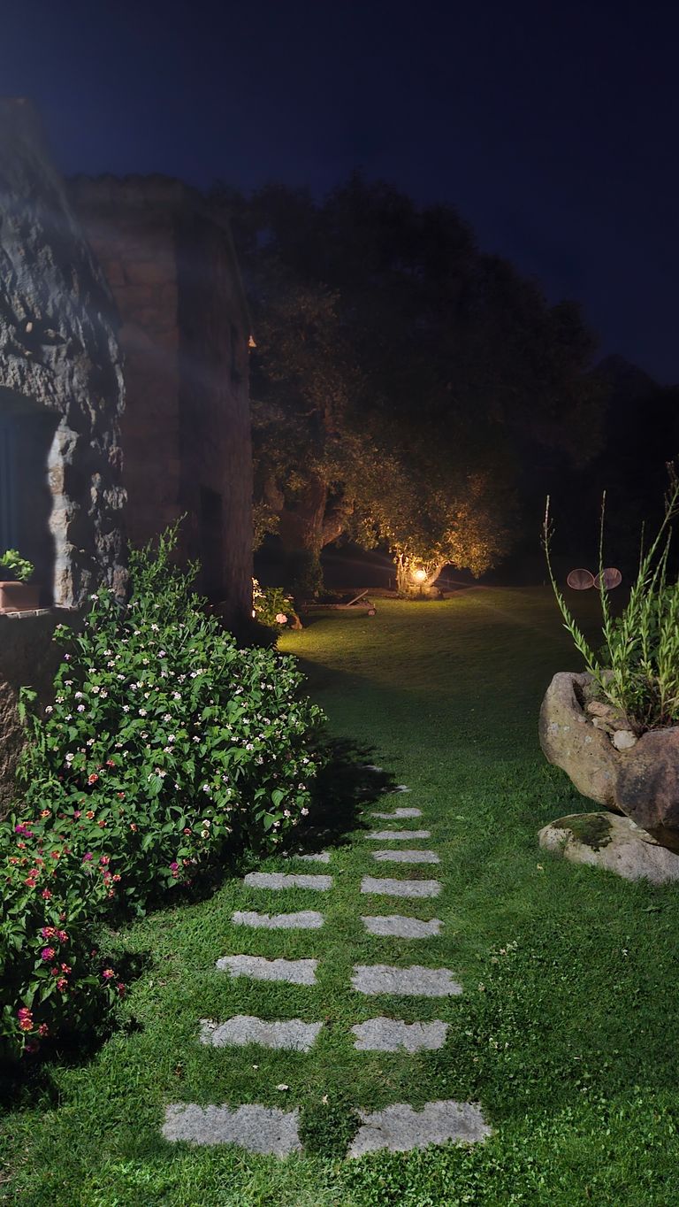 Ein beleuchteter Gartenweg aus Steinplatten führt an einem steinernen Haus und blühenden Büschen vorbei zu einem Baum, der von einer Lampe in der Nacht erhellt wird.