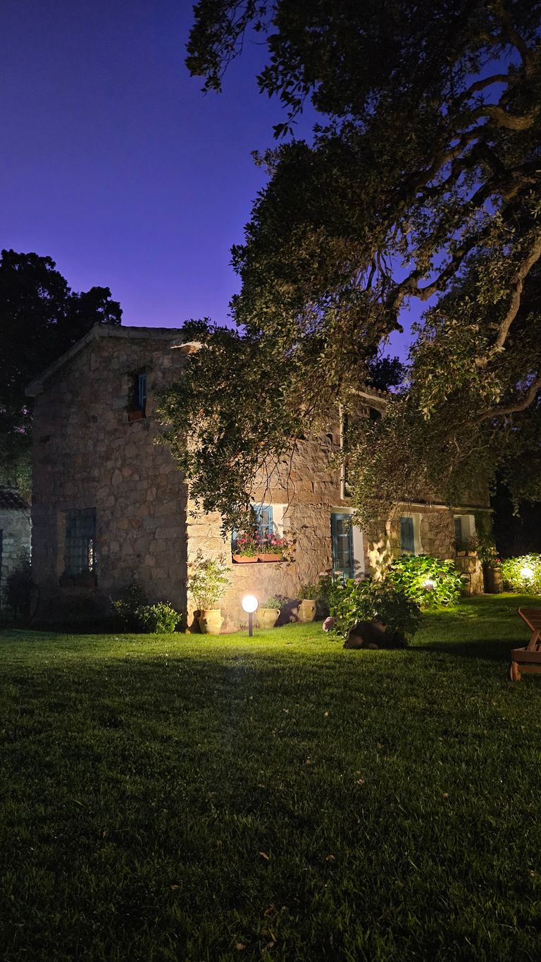 Stein gebautes Haus bei Nacht mit beleuchtetem Garten und großem Baum im Vordergrund unter einem dunkelblauen Himmel.