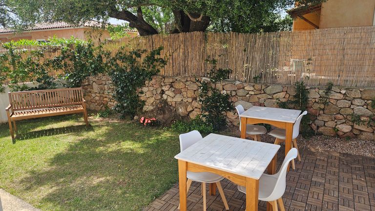 Kleiner Gartenbereich mit zwei weißen Tischen und Stühlen auf einem gepflasterten Bereich, daneben eine Holzbank auf einer Grasfläche vor einer Steinmauer und einem Sichtschutz aus Bambus.
