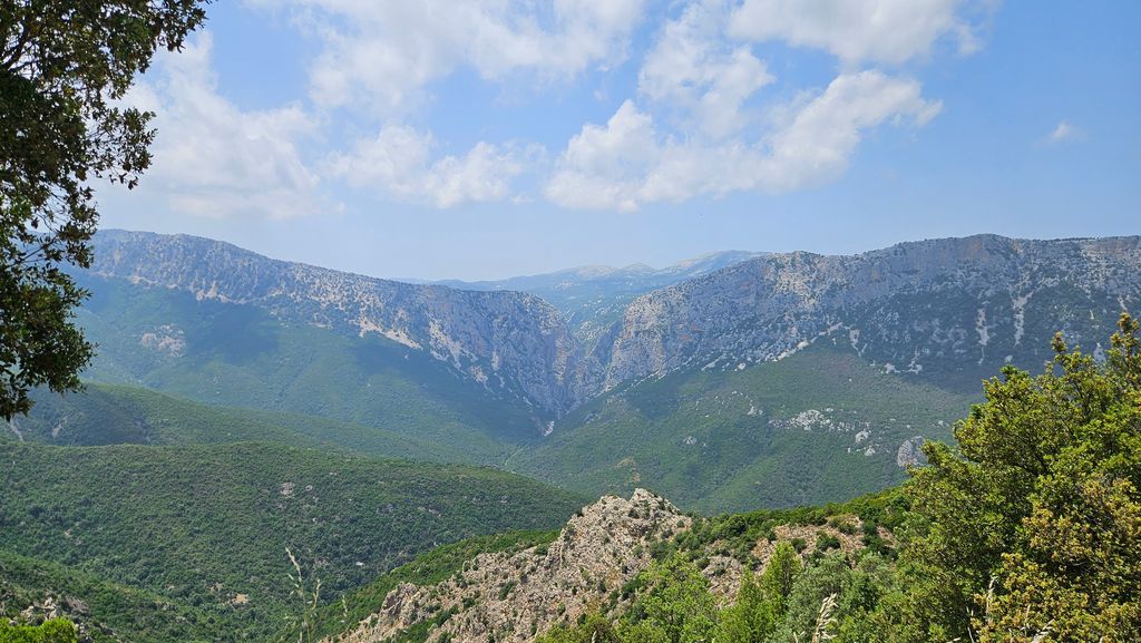 Blick auf eine bewaldete Berglandschaft mit steilen Klippen unter einem bewölkten blauen Himmel.