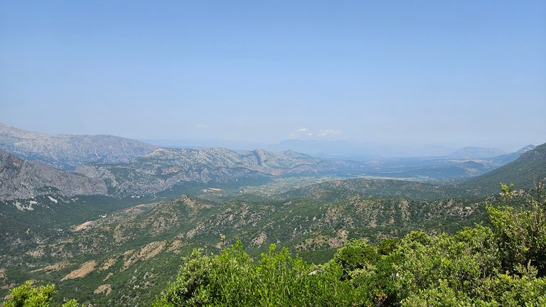 Berglandschaft mit grünen Hügeln und bewaldeten Flächen unter klarem blauem Himmel.