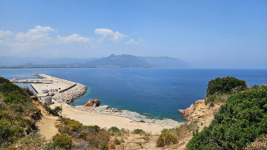 Blick auf das blaue Meer mit einem langen Hafenpier und felsigem Ufer im Vordergrund sowie Bergen im Hintergrund unter klarem Himmel.