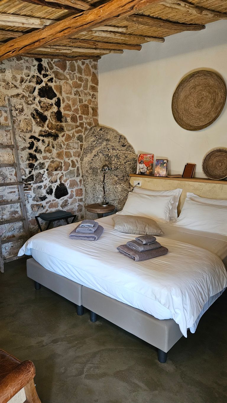 Schlafzimmer mit Doppelbett, weißen Bettlaken, gefalteten Handtüchern und Kissen, steinerner Wand, Holzbalkendecke und dekorativen Wandbehängen.