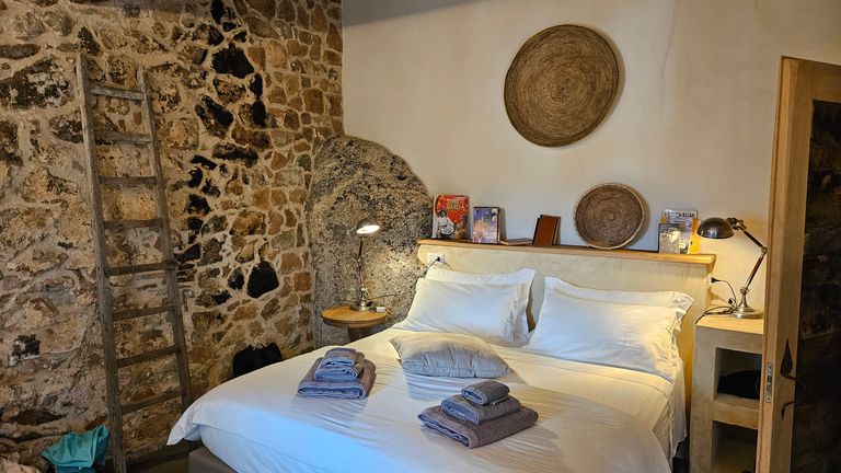 Gemütliches Schlafzimmer mit Doppelbett, weißen Bettwäsche, zwei Stapeln gefalteter Handtücher und Wandleitern an einer Steinmauer.