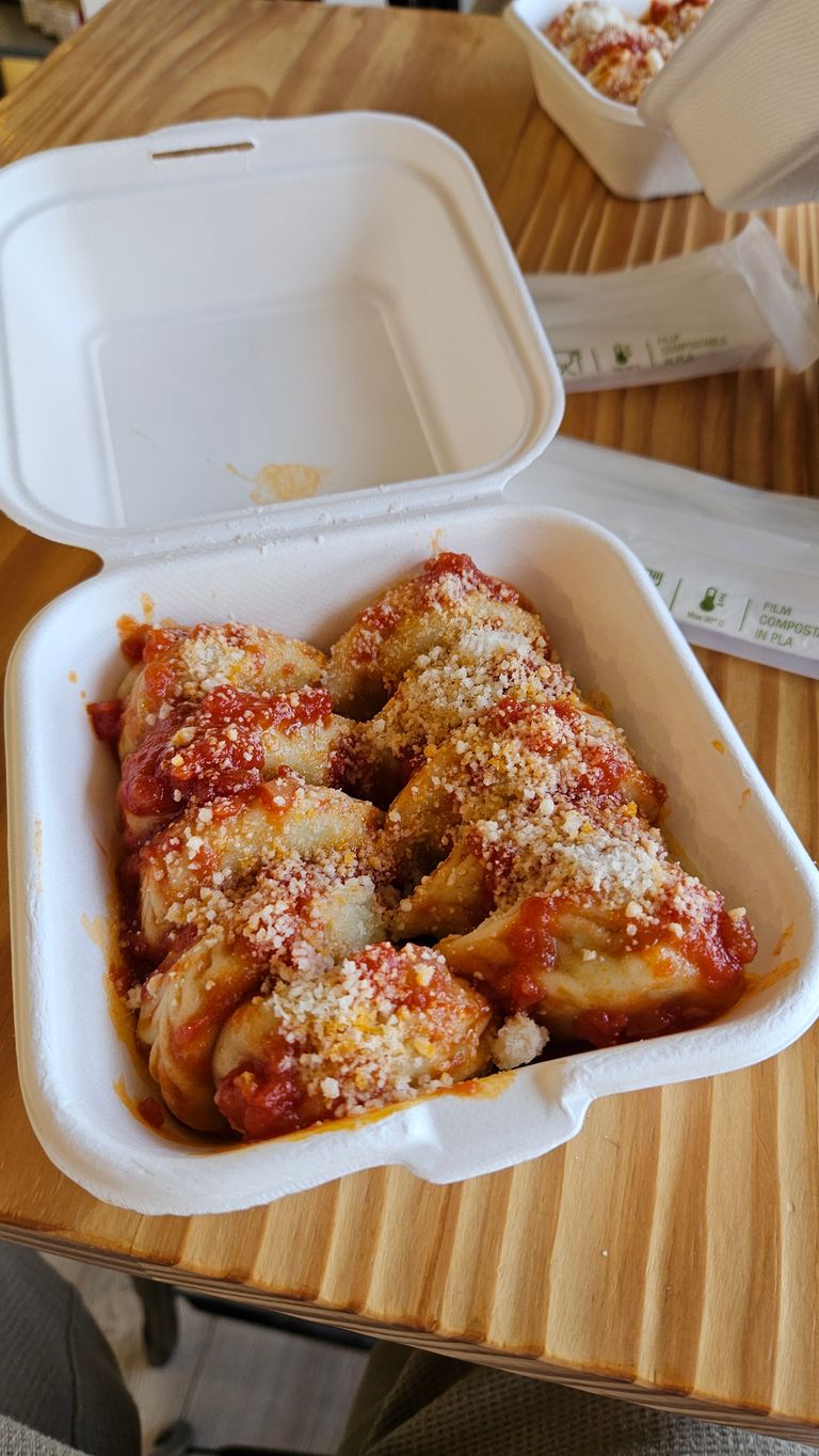 Offene weiße kompostierbare Verpackung auf einem Holztisch mit mehreren mit Tomatensauce und geriebenem Käse bedeckten gefüllten Teigtaschen.