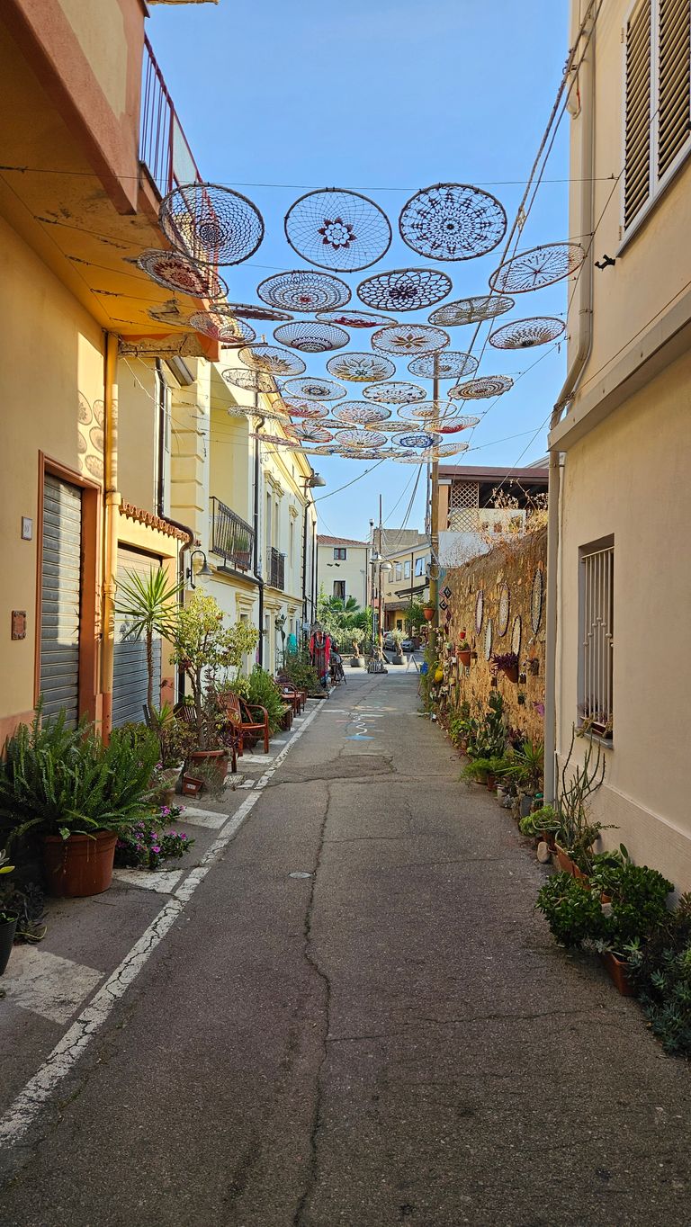 Enge Straße mit gelben und cremefarbenen Gebäuden, darüber hängende runde gehäkelte Dekorationen, viele Pflanzen in Blumentöpfen entlang der Straße.
