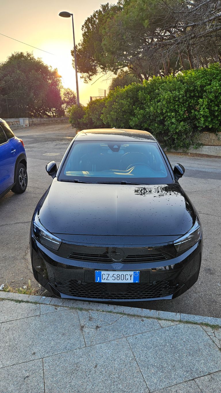 Ein schwarzes Auto mit italienischem Nummernschild, das an einer Straße neben einer grünen Hecke und Bäumen geparkt ist, bei Sonnenuntergang mit Straßenlaterne im Hintergrund.