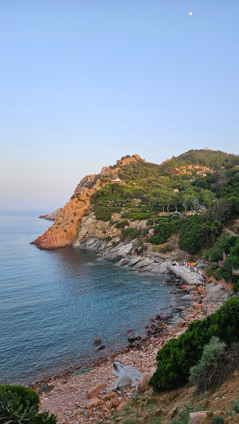 Küstenlinie mit felsigem Ufer, grün bewachsenen Hügeln und einer kleinen Promenade mit weißen Sonnenschirmen und Stühlen am Meer unter klarem Himmel mit sichtbarem Mond.