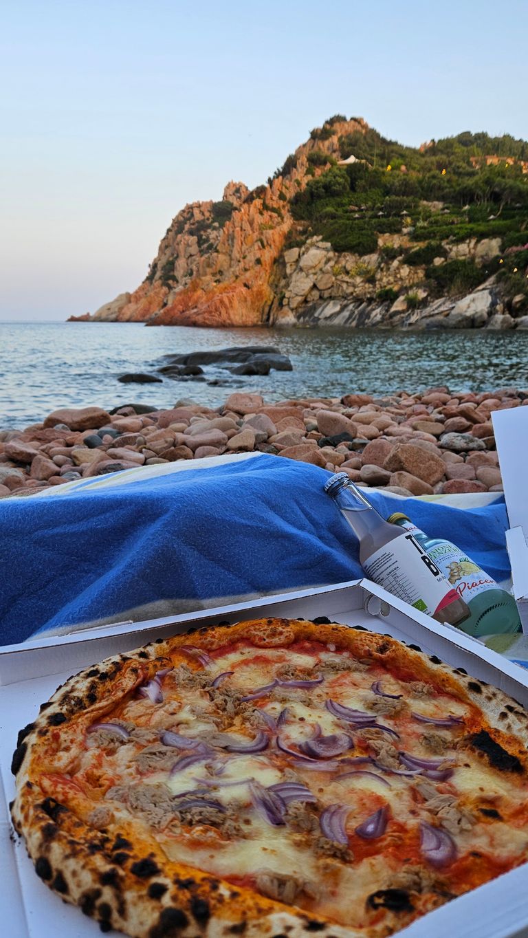 Eine Pizza mit Zwiebeln und Thunfisch in einer Pizzaschachtel am felsigen Strand mit Meer und bewaldeten Klippen im Hintergrund bei Sonnenuntergang.