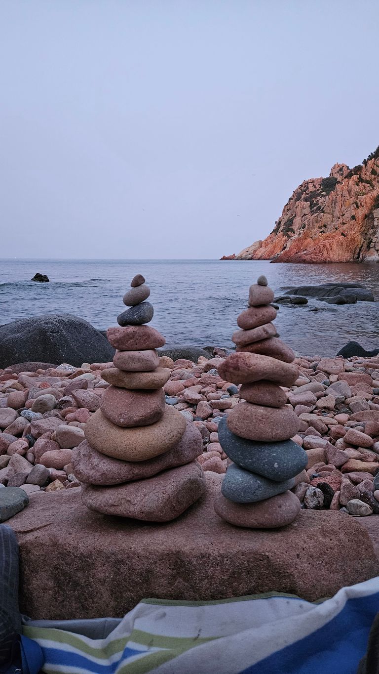 Zwei kunstvoll gestapelte Steinhaufen am felsigen Strand mit ruhigem Meer und einem Felsen im Hintergrund bei bewölktem Himmel.