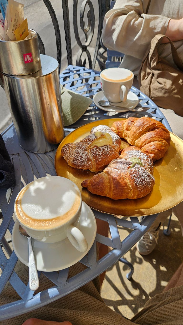 Zwei Tassen Cappuccino und ein goldener Teller mit drei Croissants auf einem blauen Metalltisch im Freien, eine Person sitzt daneben mit brauner Jacke und Rucksack.