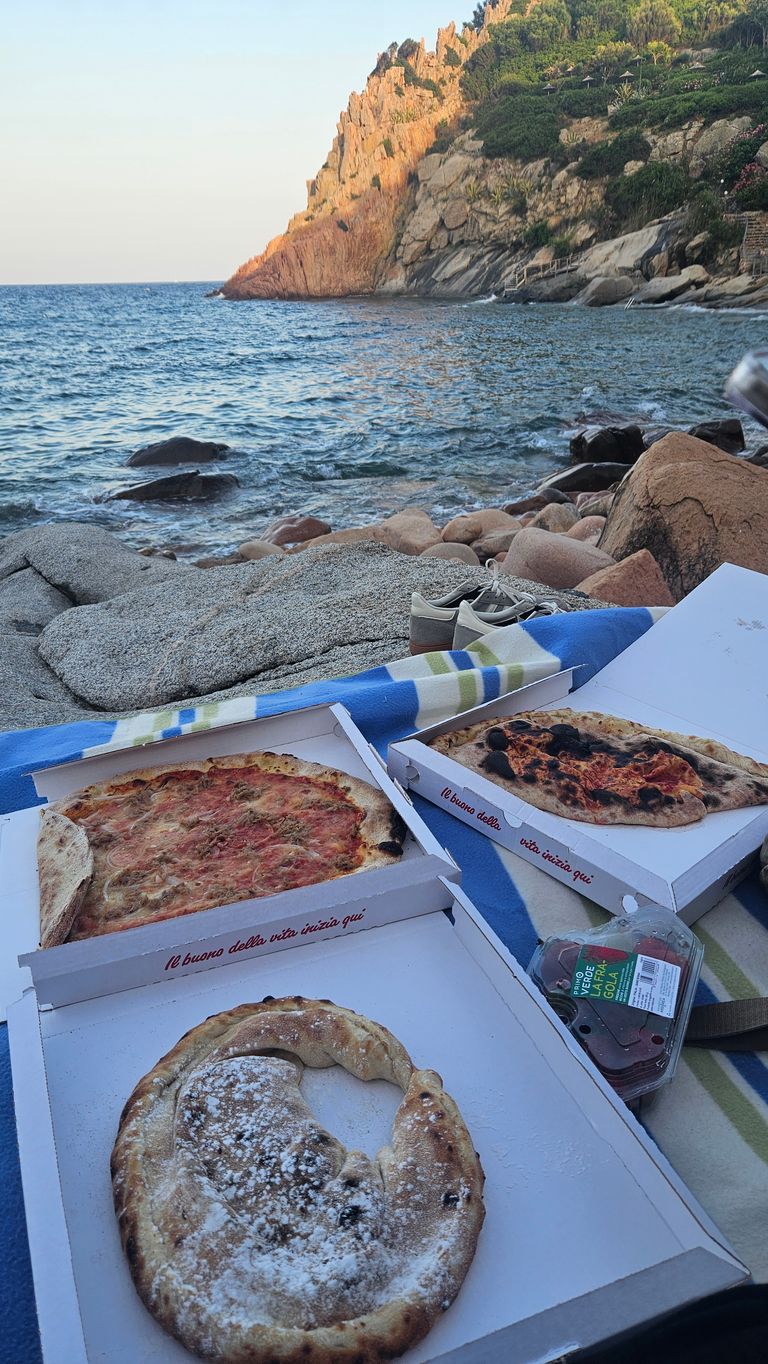 Drei geöffnete Pizzaschachteln mit unterschiedlichen Pizzen liegen auf einer Decke am felsigen Strand neben dem Meer, im Hintergrund ein bewachsener Felsvorsprung bei Sonnenuntergang.