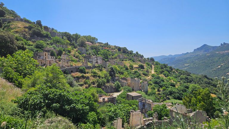 Verlassene, überwucherte Gebäude auf einem grünen Hügel mit Bergen im Hintergrund unter klarem blauem Himmel.