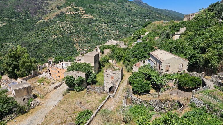 Verlassene und teilweise verfallene Gebäude an einem Berghang umgeben von grünen Bäumen und Bergen bei sonnigem Wetter.