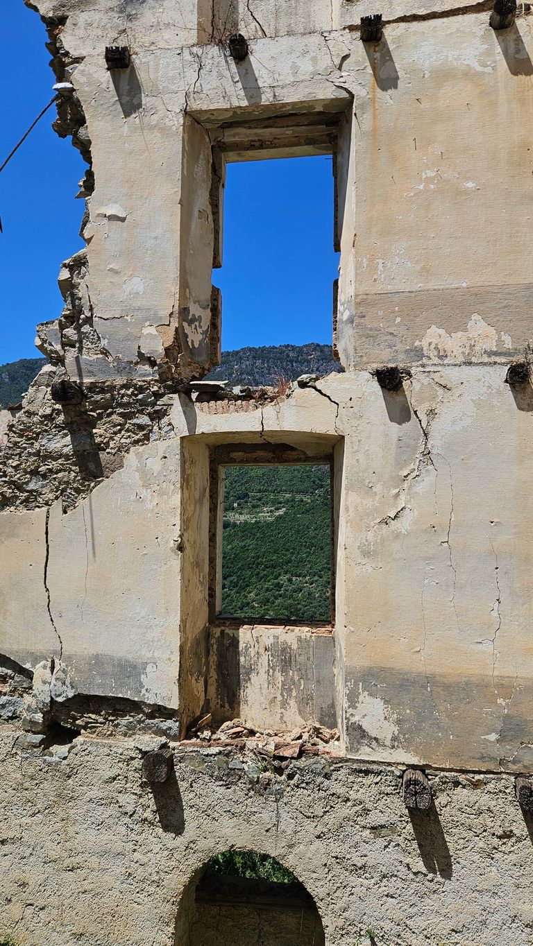 Ruinenmauer mit zwei rechteckigen Fenstern, durch die ein klarer blauer Himmel und bewaldete Hügel sichtbar sind.