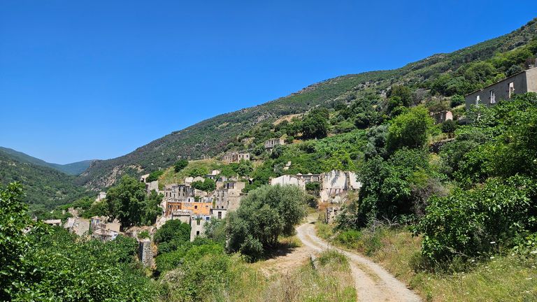 Ein kleiner Weg führt durch eine grüne, bewachsene Hügellandschaft mit verstreuten alten Häusern unter klarem blauem Himmel.