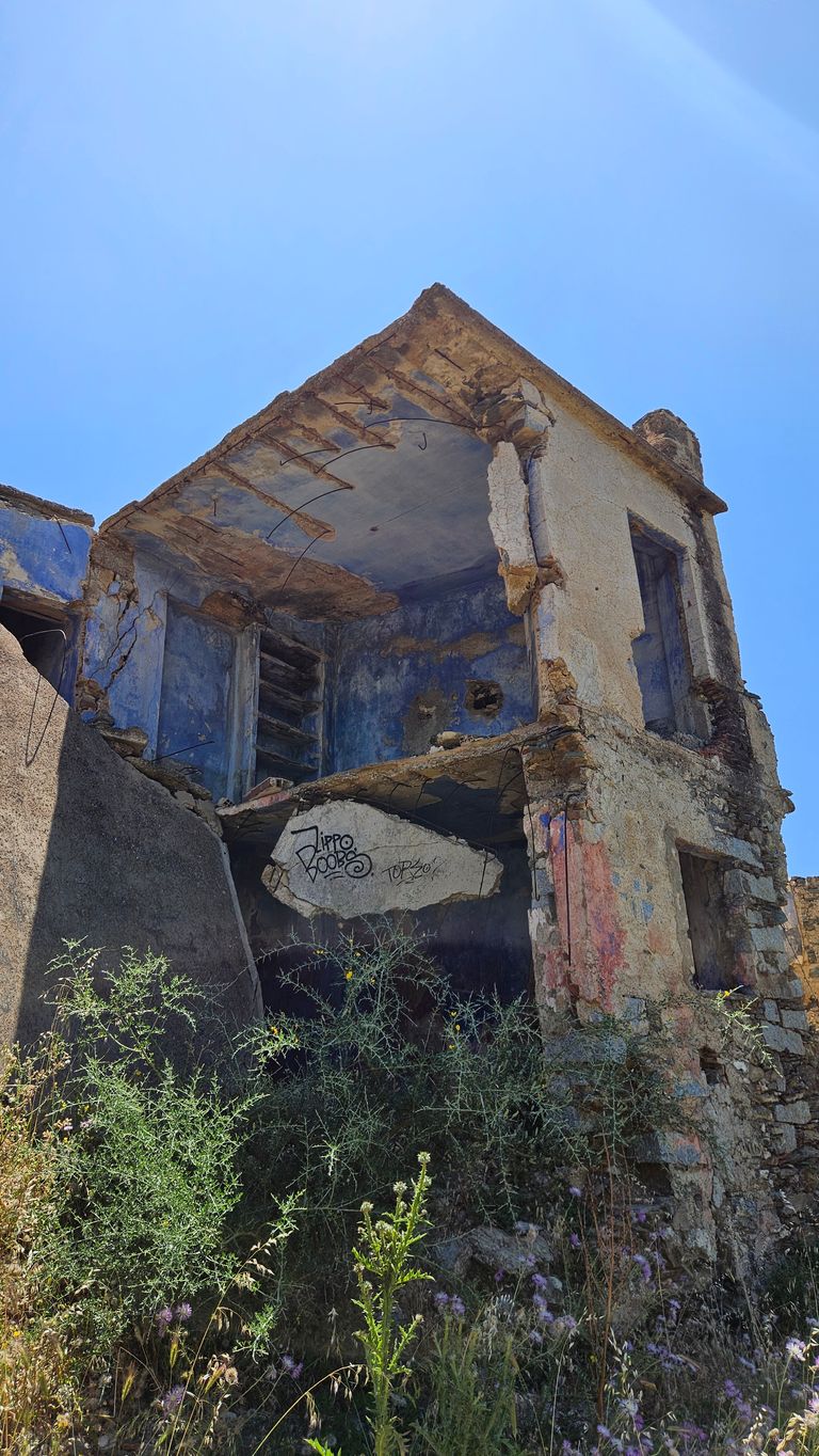 Ruinen eines zweistöckigen Gebäudes mit freigelegten inneren Wänden und einer blauen Farbgebung im Inneren, umgeben von wildwüchsigen Pflanzen unter einem klaren blauen Himmel.