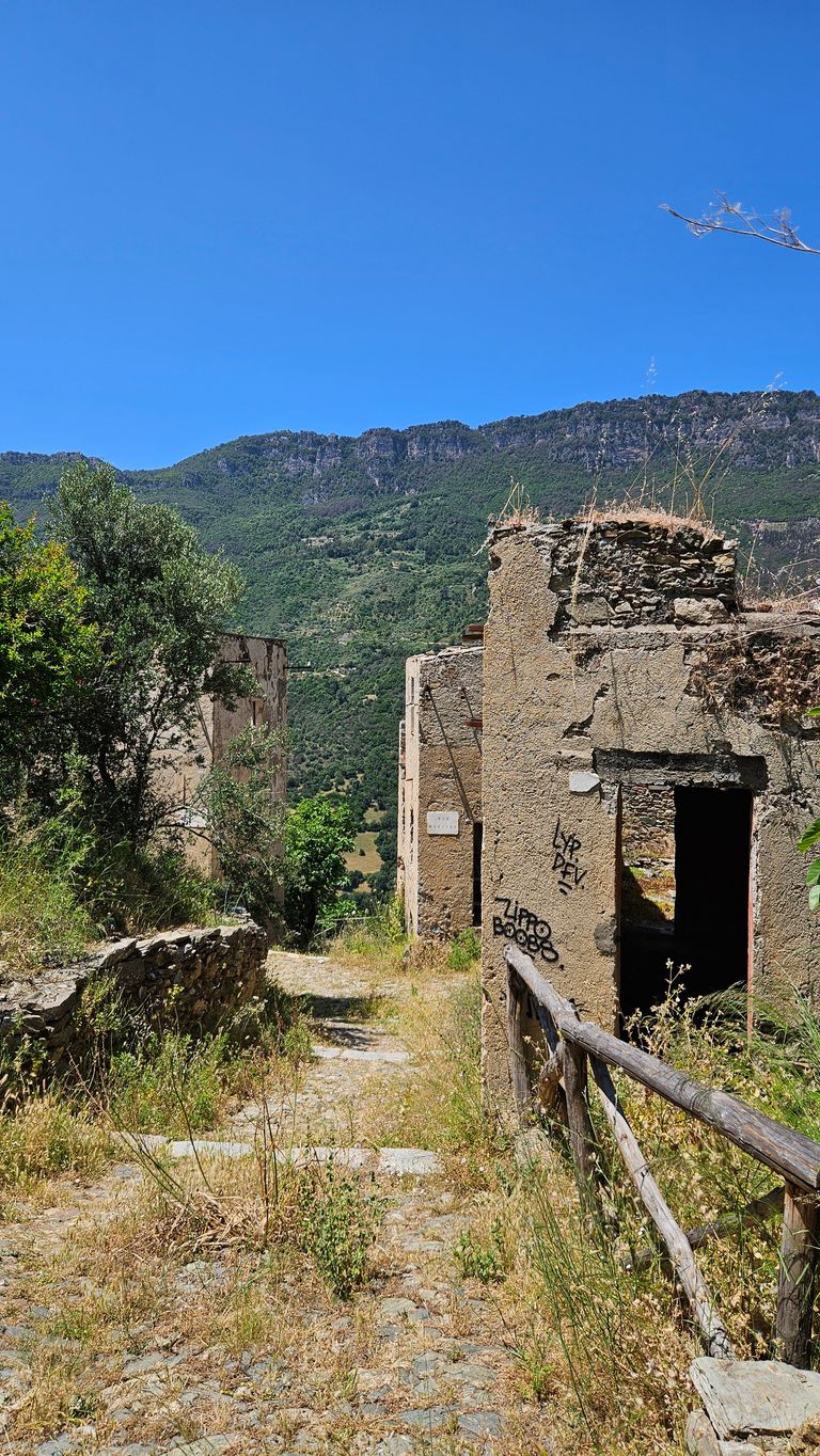 Verlassener, verfallener Steinhof mit einer Steinmauer und Gestrüpp am Weg, im Hintergrund bewaldete Berge unter klarem blauem Himmel.